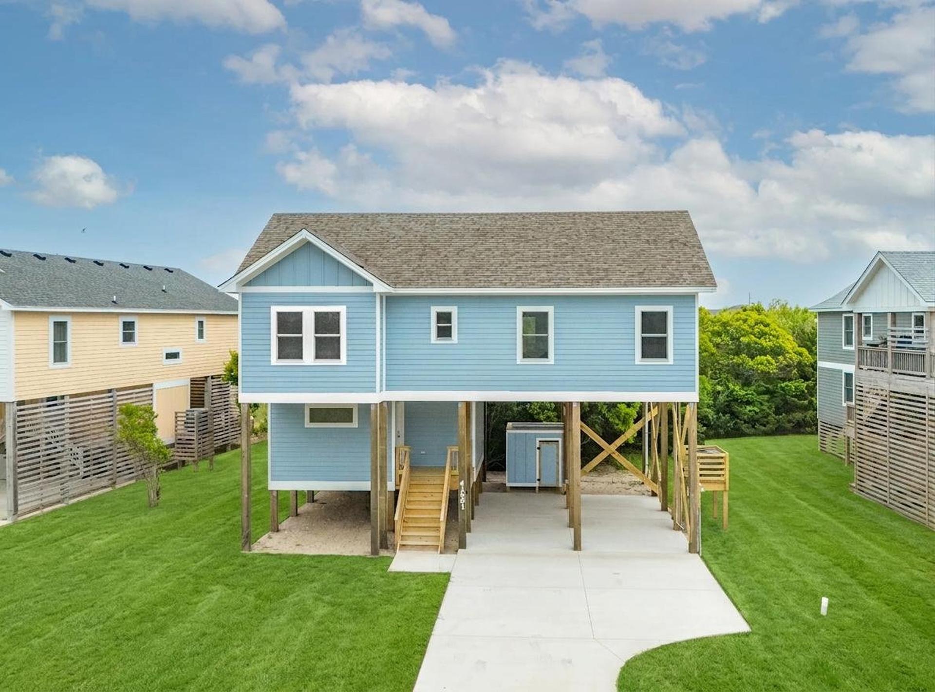 A blue raised beach house with a green lawn and a concrete driveway